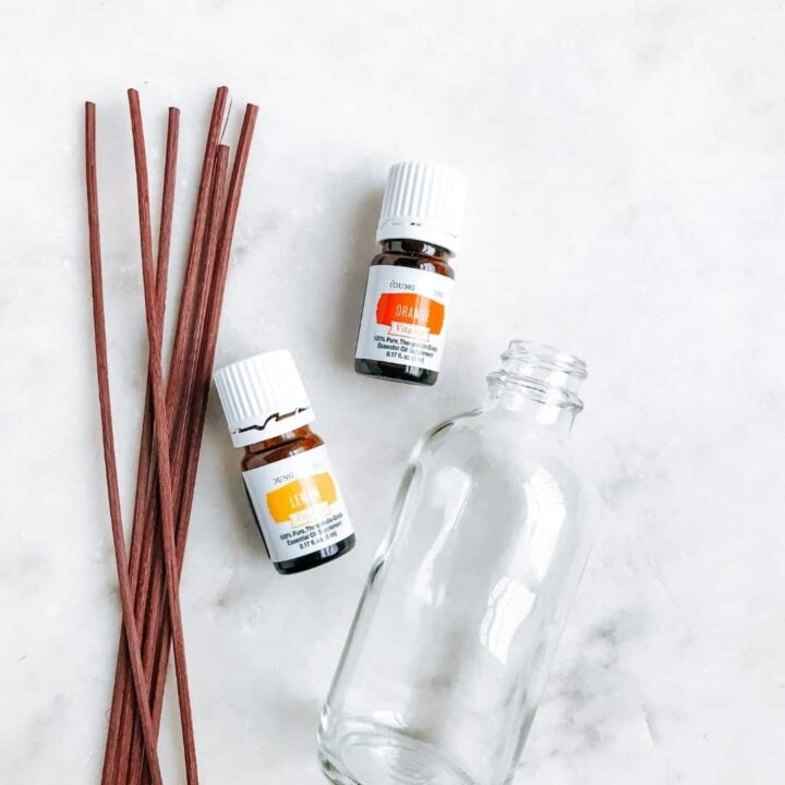 glass bottle laying next to brown reed diffusers and bottles of lemon and orange essential oils. Showing tools needed to make a reed diffuser